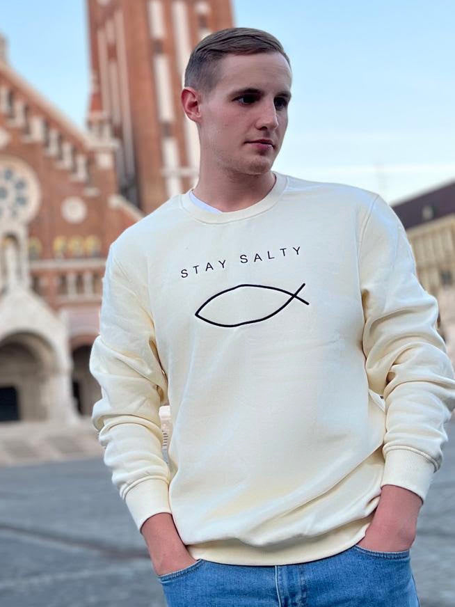 Cream Stay Salty embroidered Christian crewneck with the ichthys fish, worn by a young man in front of the twin towers of the Votive Church in Szeged, Hungary