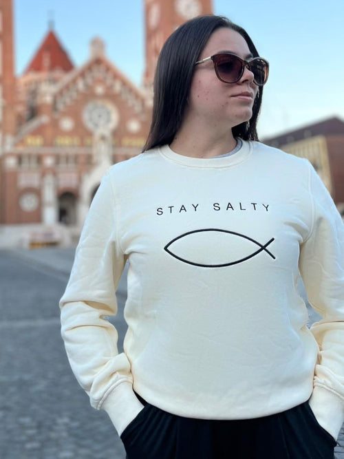 Woman in sunglasses wearing the cream Stay Salty embroidered Christian crewneck in front of the Szeged Votive Church towers, Hungary